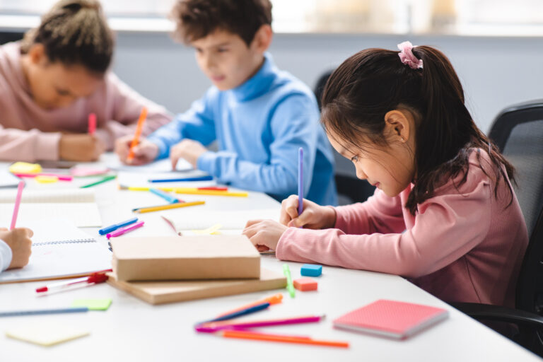 Portrait of small asian girl sitting at table in classroom at primary school or kindergarten, drawing in notebook with colorful pencils. Reopening and return back to school after lockdown